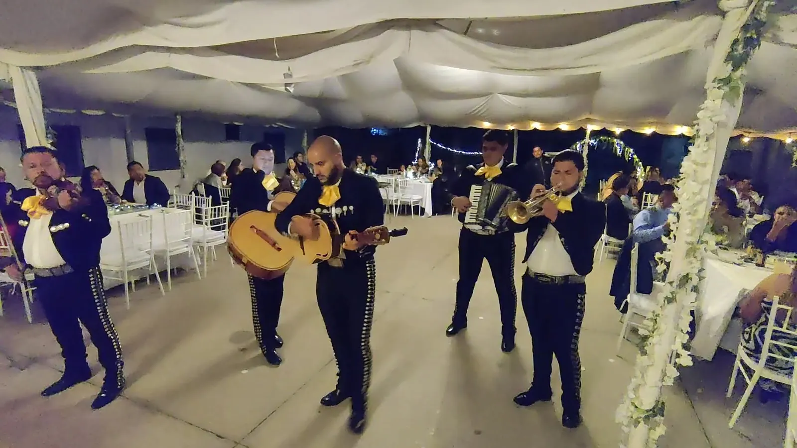 Los músicos de Mariachi Jalisco Es Tradición, en traje de charro negro con botonadura de plata y moño amarillo, tocando violín, guitarrón, vihuela, acordeón y trompeta en un evento de gala.